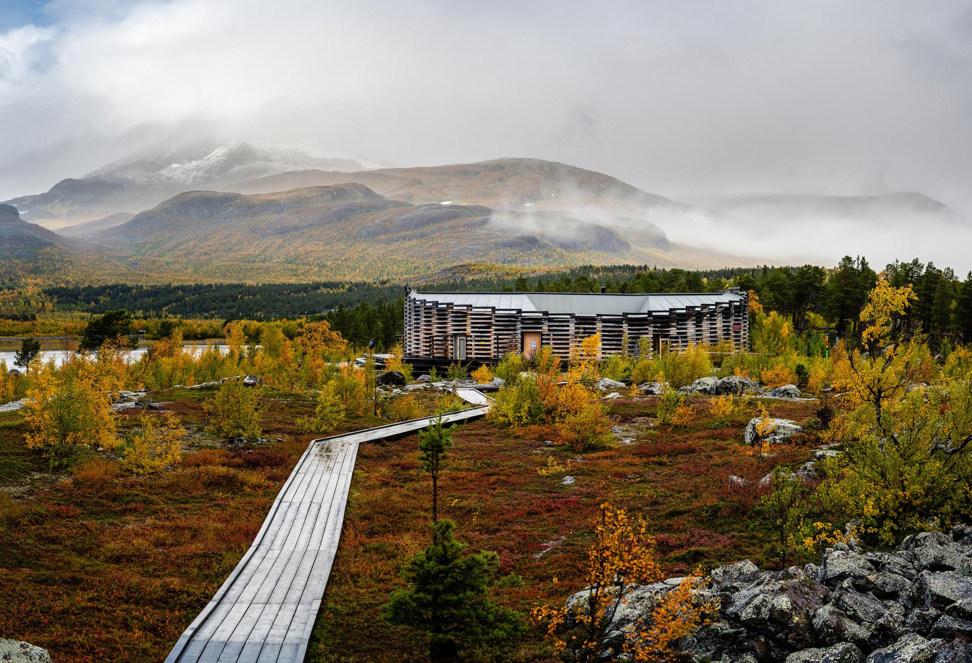 Ein Holzsteg führt zum Gebäude des Naturum Laponia. Hinter dem Gebäude befinden sich Berge, die leicht von Nebel bedeckt sind.