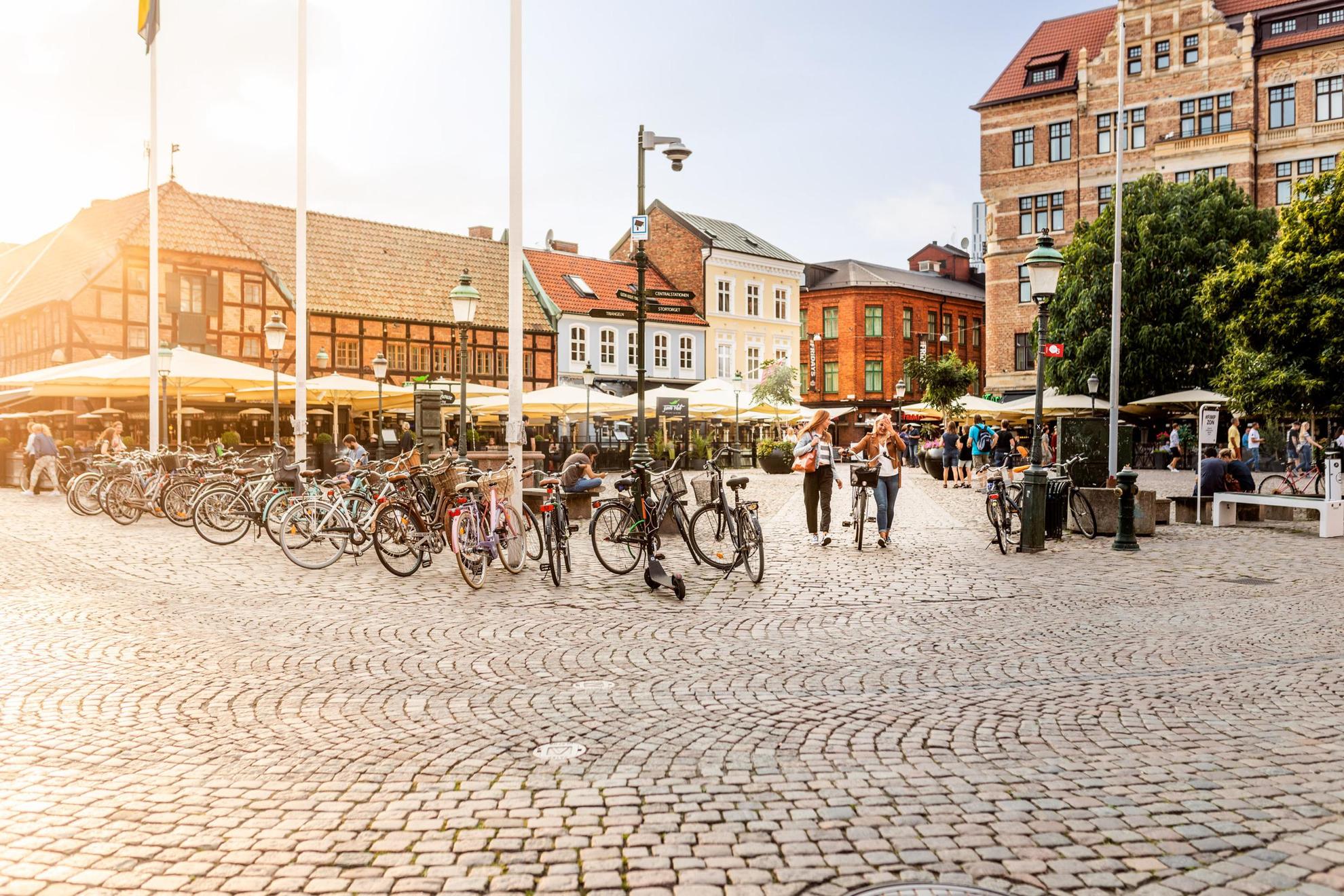 Ein Platz in einer Altstadt, Backstein- und Steinhäuser an der Seite. Straßen-Restaurants mit Sonnenschirmen. Auf dem Platz stehen Menschen, die auf Bänken sitzen, und mehrere Fahrräder sind geparkt.