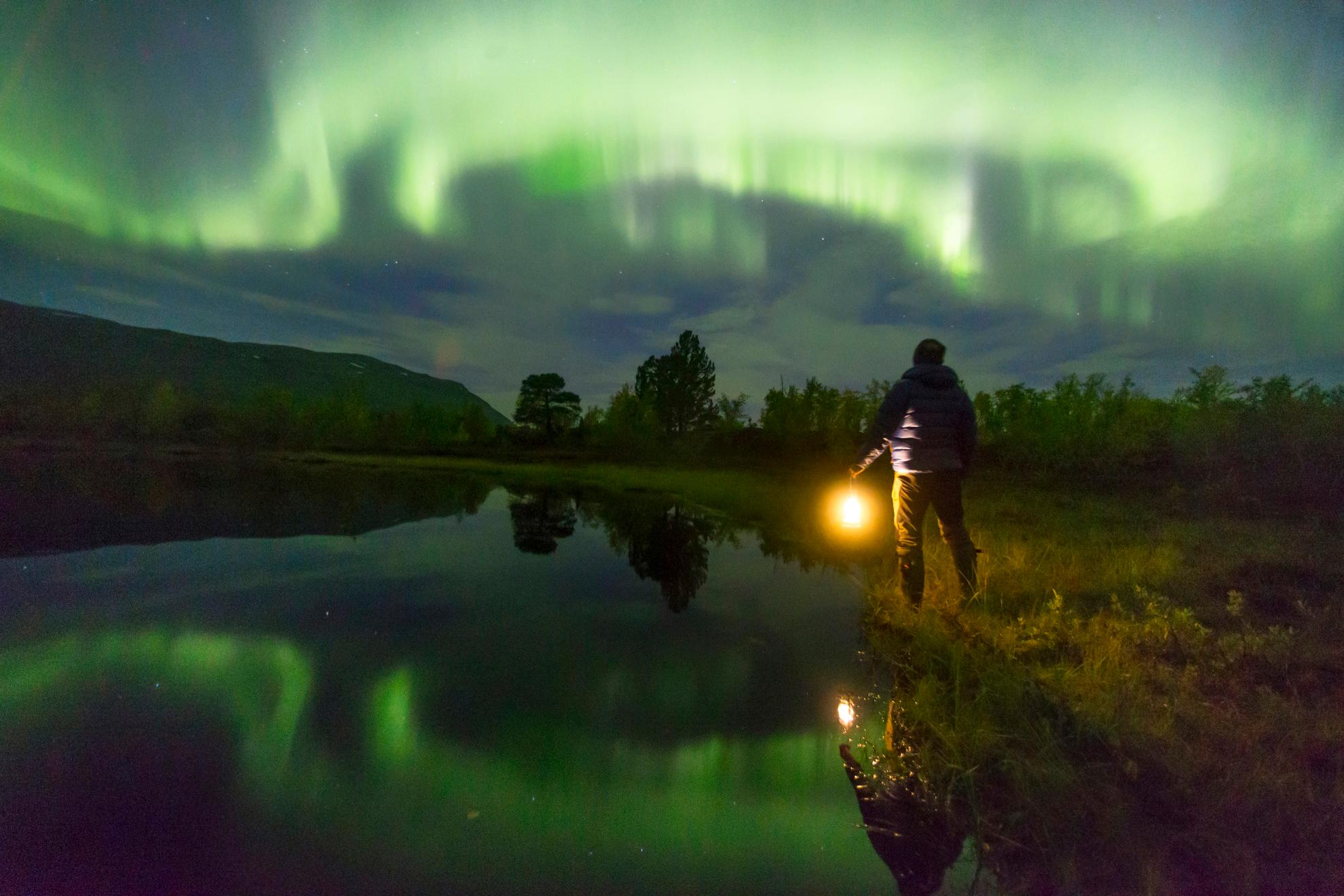 Eine Person mit einer Lampe blickt auf das grüne Nordlicht am Himmel. Die Nordlichter spiegeln sich in dem See neben der Person.
