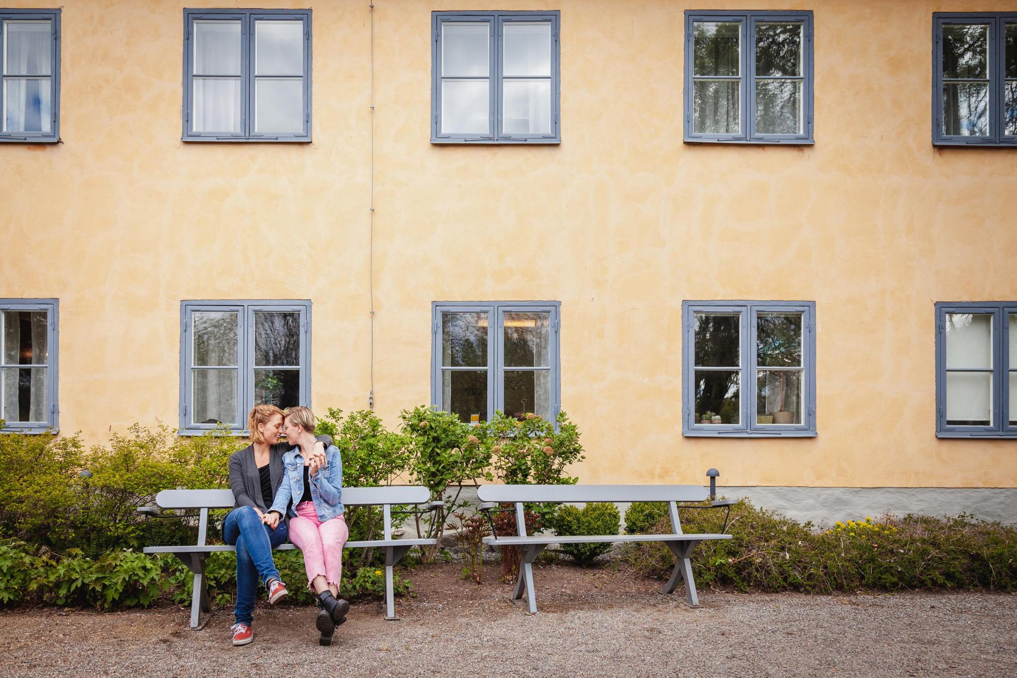 Zwei Frauen küssen sich und halten sich an den Händen, während sie auf einer Parkbank vor einem Haus sitzen.