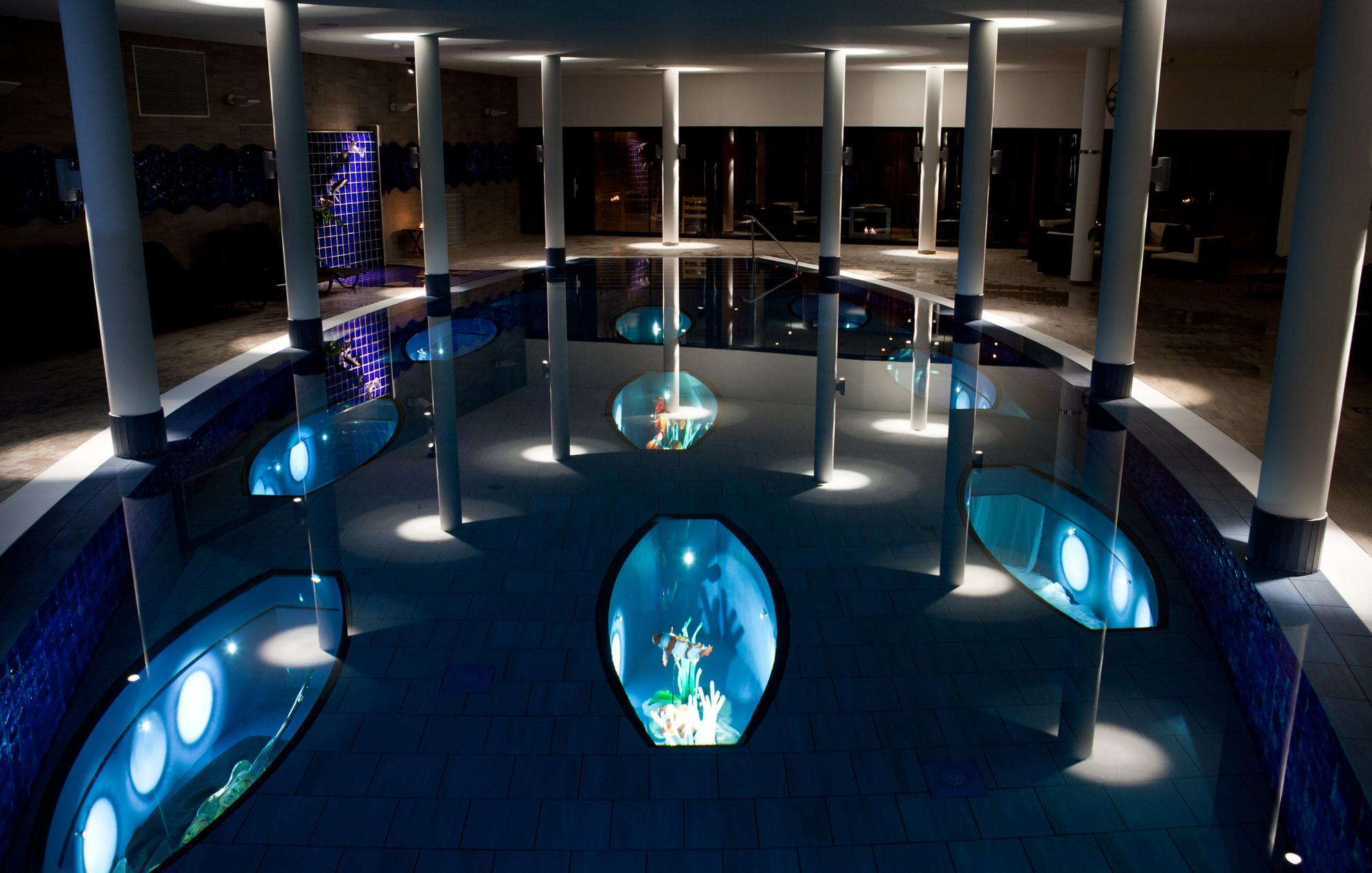 Ein Hallenbad, in dem das Licht gedimmt ist. Im Becken befinden sich beleuchtete Kunstausstellungen mit Glasfischen.