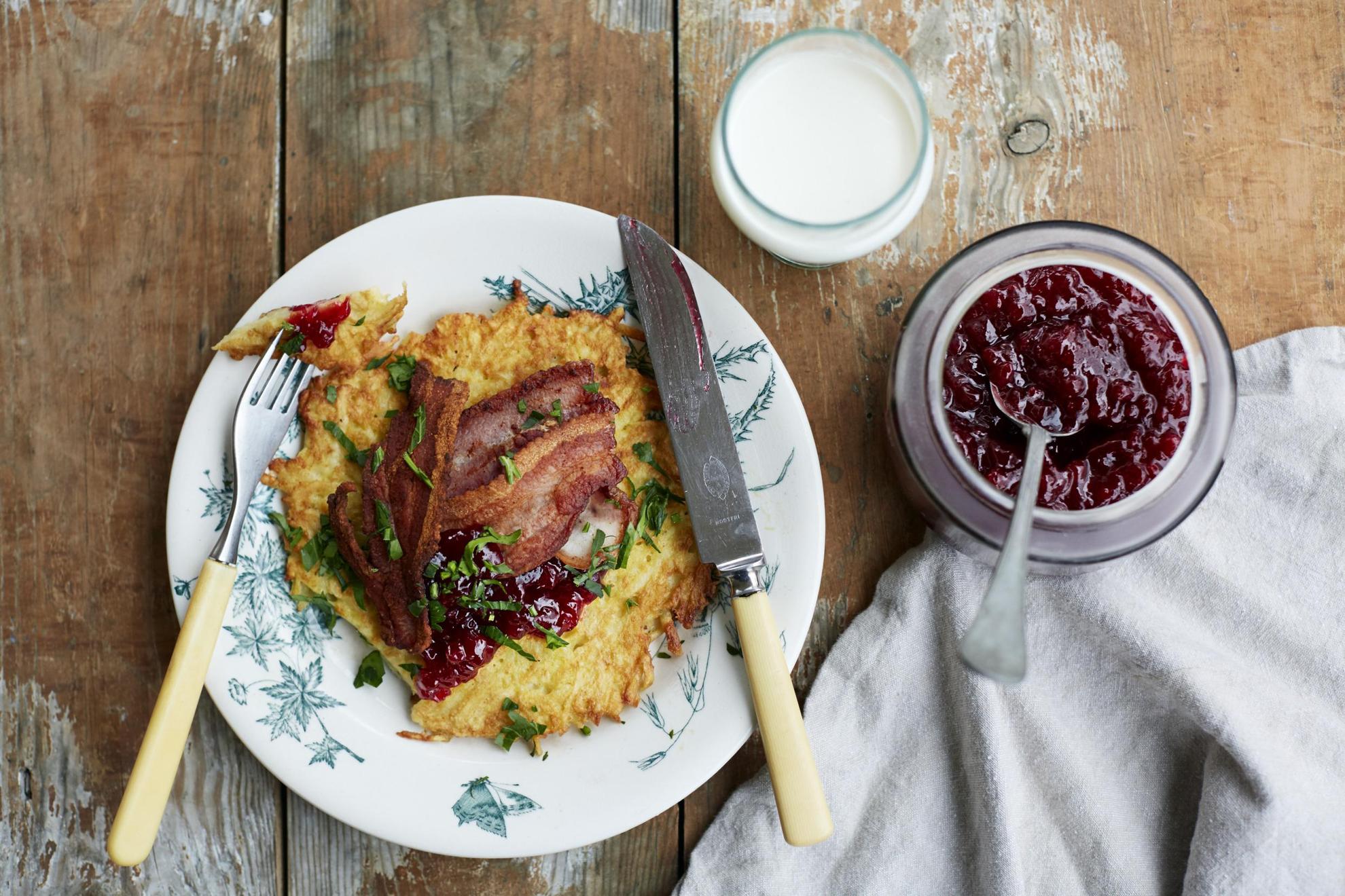Seen from above, a wooden table is set with a plate of potato pancakes and bacon, a jar of lingonberry jam and a glass of milk.