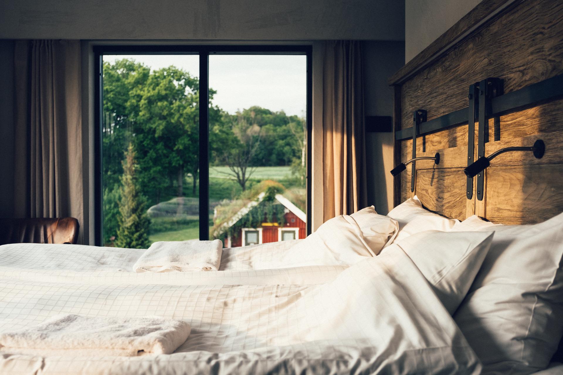 Ein Doppelbett in einem Hotelzimmer mit Fenstern, die den Blick auf eine rote Hütte und einen Wald freigeben.
