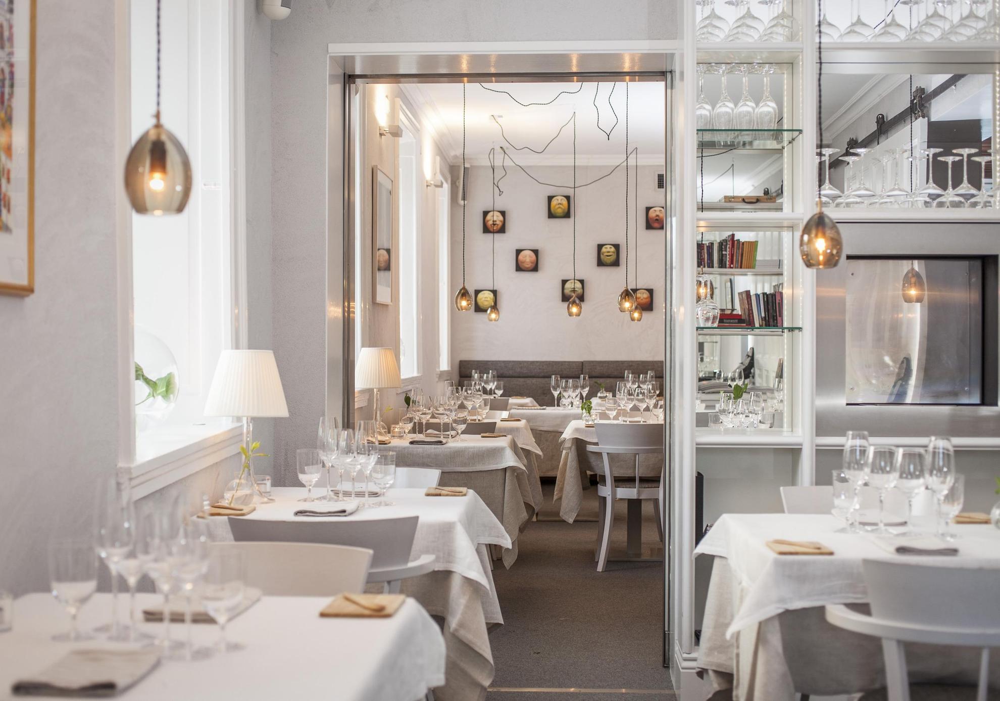 In einem Restaurant mit weißen Wänden sind die Tische mit weißen Tischdecken, Leinenservietten und Weingläsern gedeckt. Lampen hängen von der Decke.